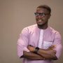 A young, handsome African man wearing a stylish purple native outfit paired with nerdy glasses. He has his arms crossed and smiles warmly while looking away, blending traditional fashion with a modern, intellectual vibe.