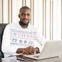 Young African man at work with laptop computer. Man working in office with striped wall, wearing patterned shirt. Male office worker using laptop, Nigeria, Africa.